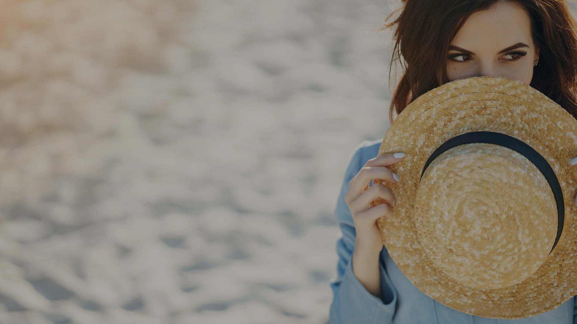 A person on the beach covering part of his face with a straw hat.