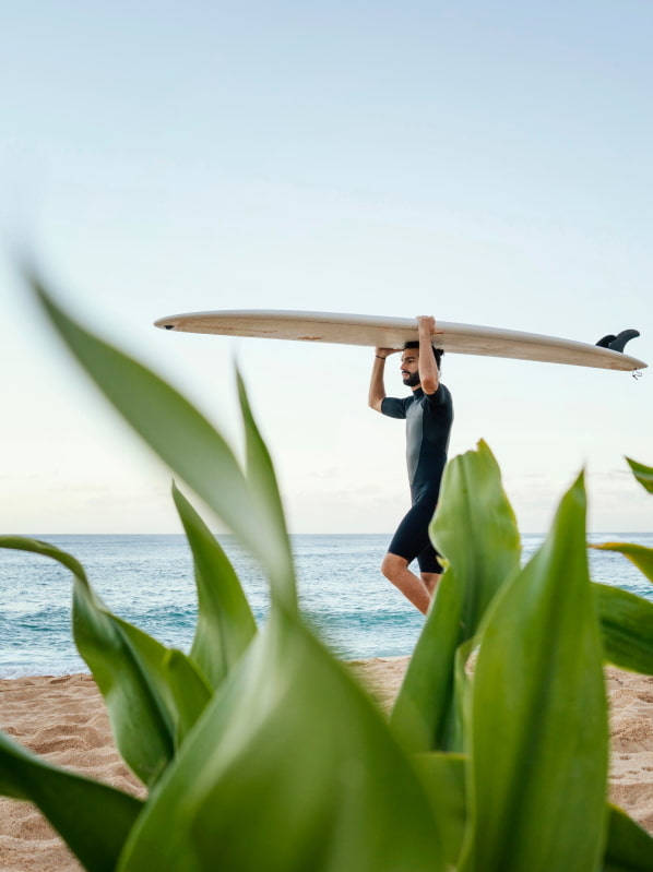 Hombre vestido en neopreno llevando sobre su cabeza una tabla de surf